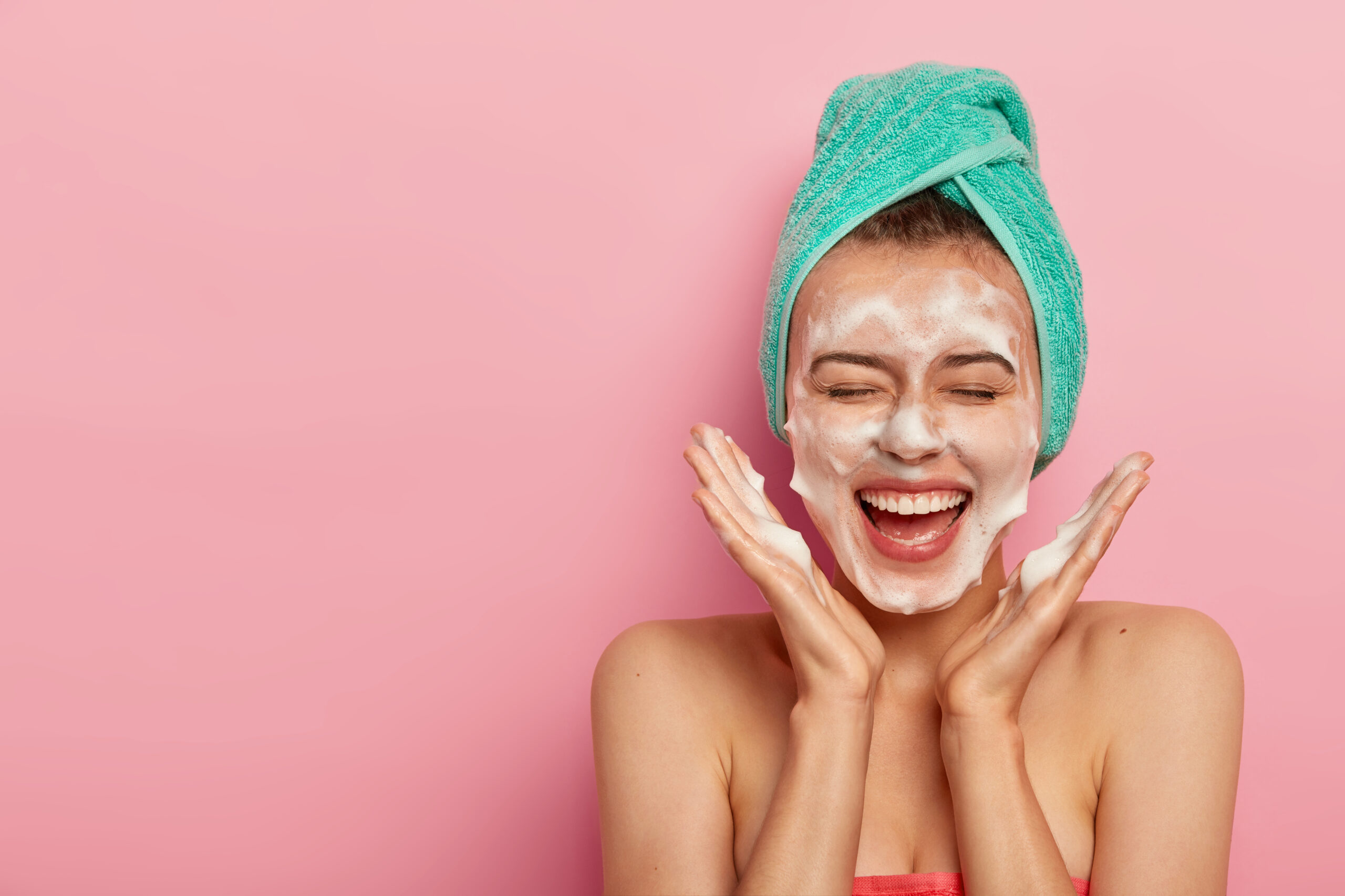 Happy joyous young girl spreads palms over face, washes face with soap, has fun in bathroom, pampers skin, wears wrapped towel on head, expresses positive emotions, poses against pink background
